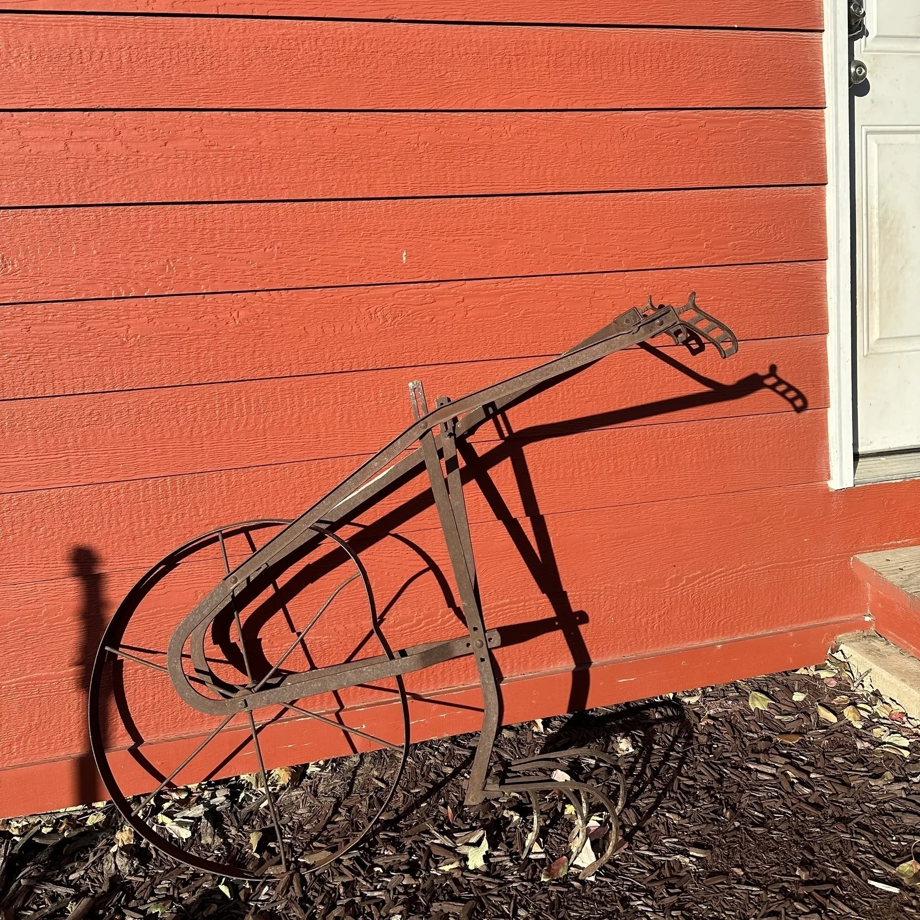 An old-fashioned, rusted cultivator is leaning against a red wooden wall with a white door nearby.
