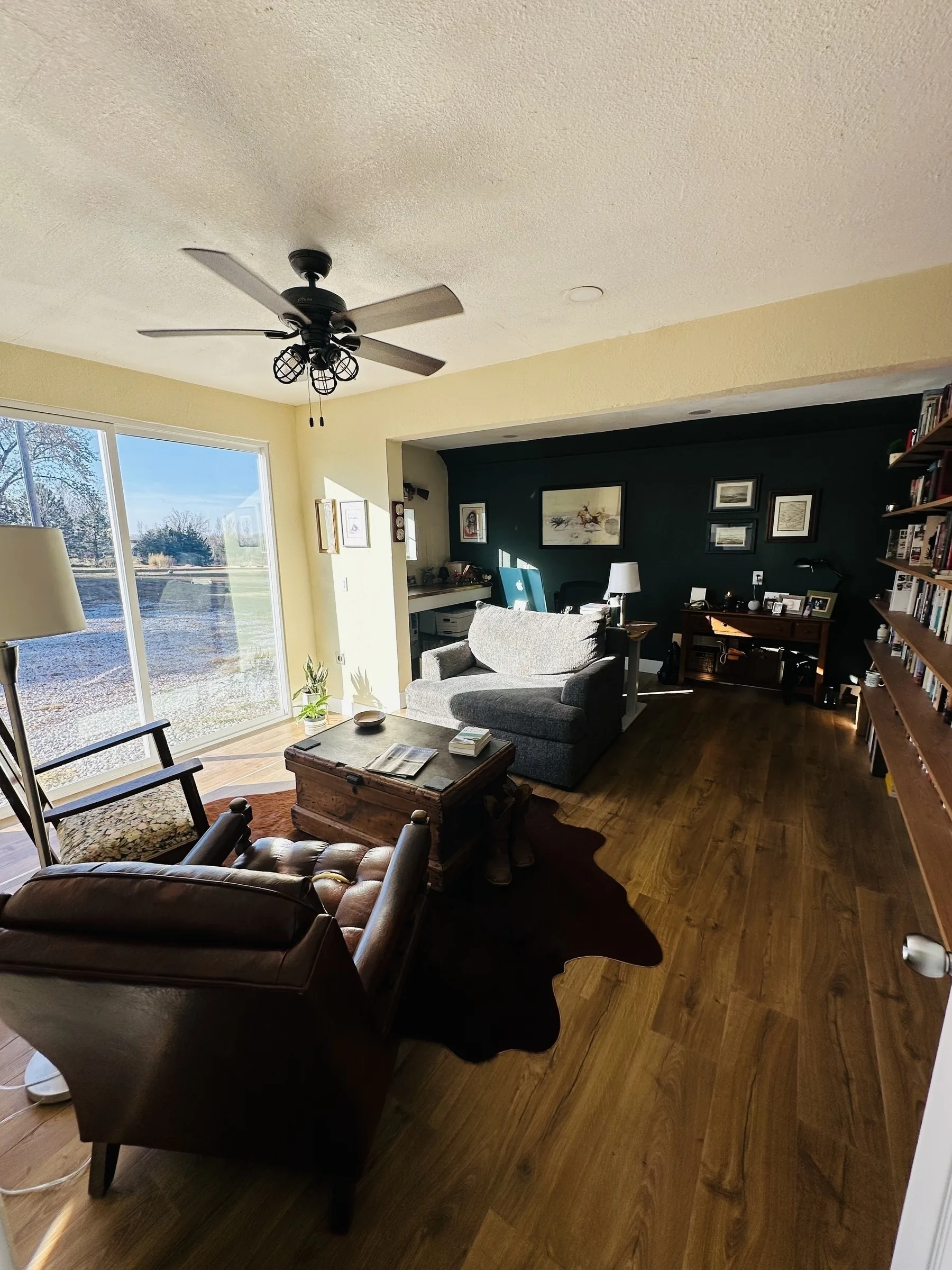 A cozy living room features a leather chair, a plush sofa, a wooden floor, a ceiling fan, and sliding glass doors leading to a patio.