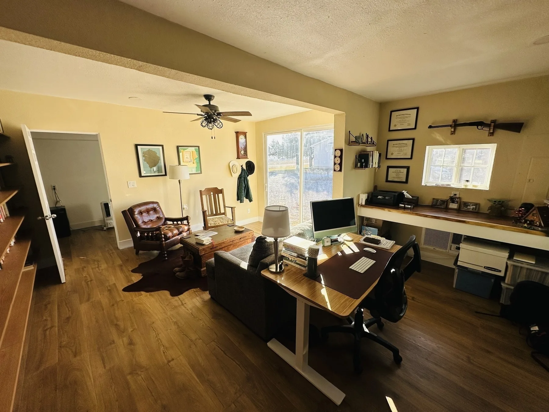 A home office features a wooden desk with a computer, a comfortable seating area, and decor such as framed pictures and certificates on the walls.