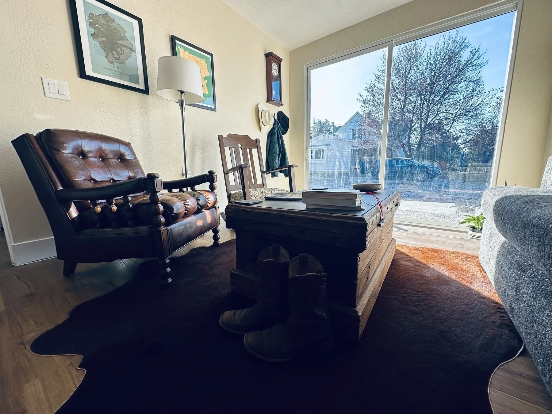 A cozy living room features a leather chair, a wooden chest as a coffee table, artwork on the walls, and large windows letting in natural light.