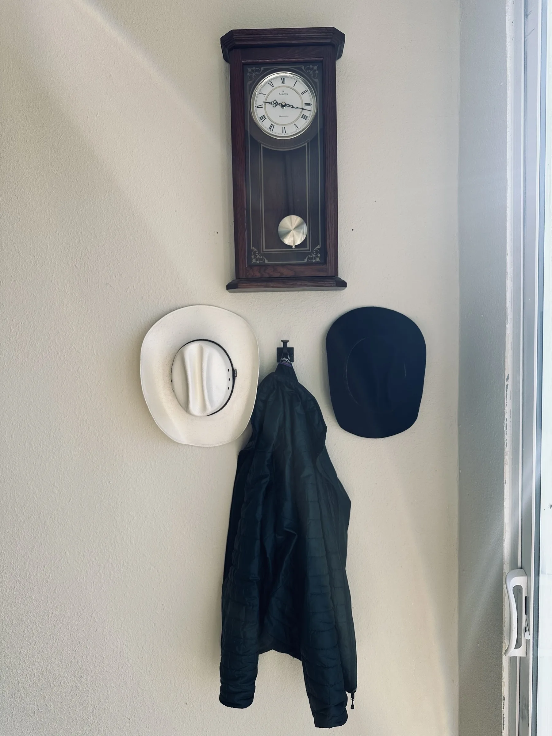A wall clock hangs above two hats and a jacket on hooks beside a window.