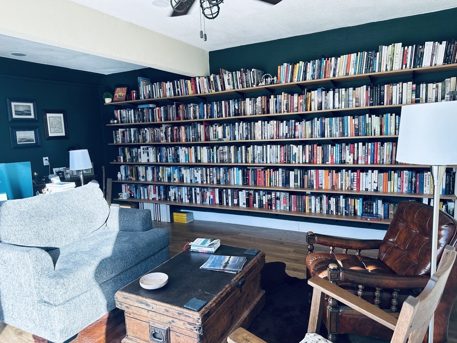 A cozy living room features a large bookshelf filled with books, a comfortable gray sofa, a brown leather chair, and a wooden coffee table.