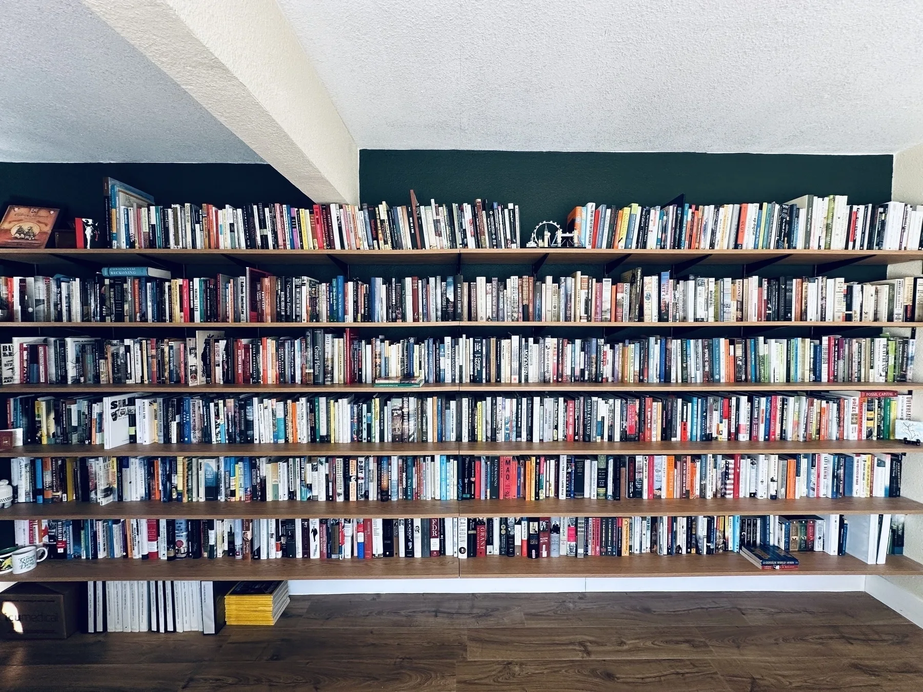 A large bookshelf filled with a diverse collection of books spans an entire wall in a room with wooden flooring.