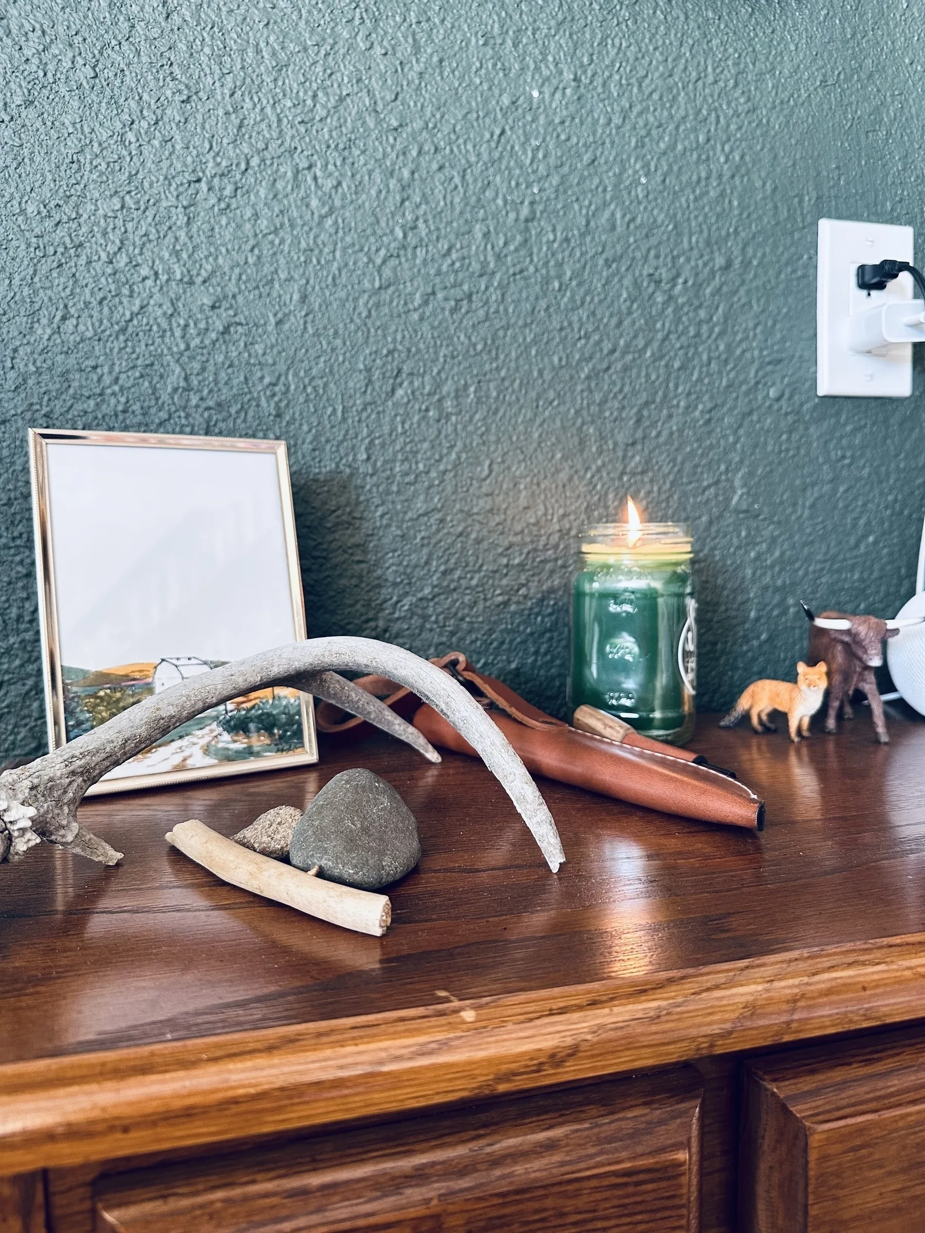 A wooden surface holds decorative items including a framed picture, a candle, small animal figurines, and natural objects like rocks and a stick against a textured green wall.