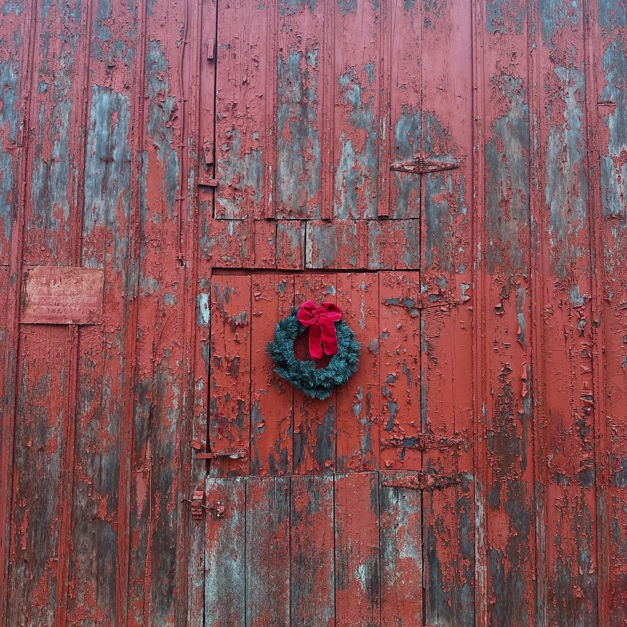 Auto-generated description: A weathered red wooden door features a green wreath with a red bow at its center.