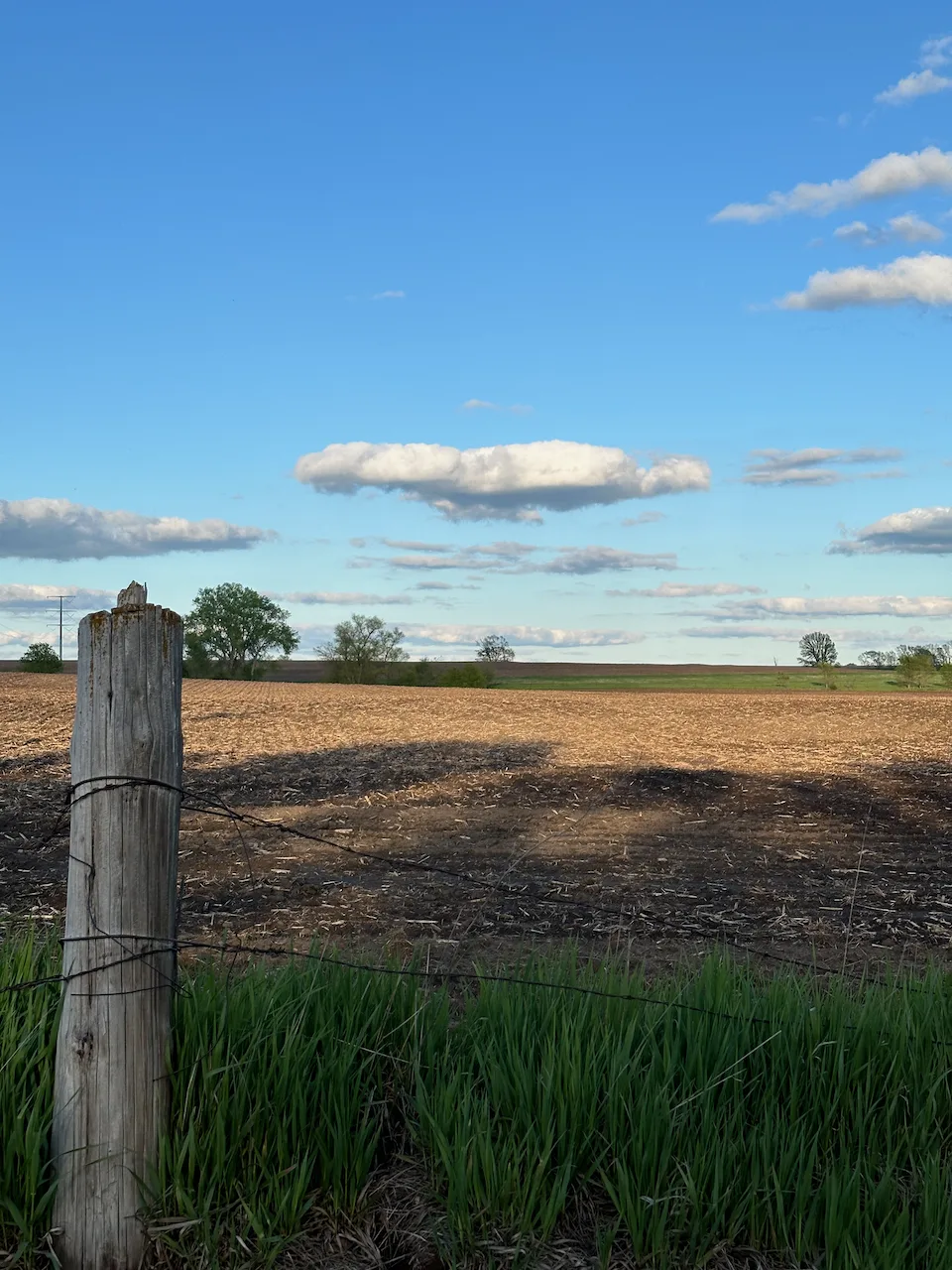 A wooden fence post stands in the foreground with green grass, while a vast, plowed field and a blue sky with scattered clouds extend into the distance.