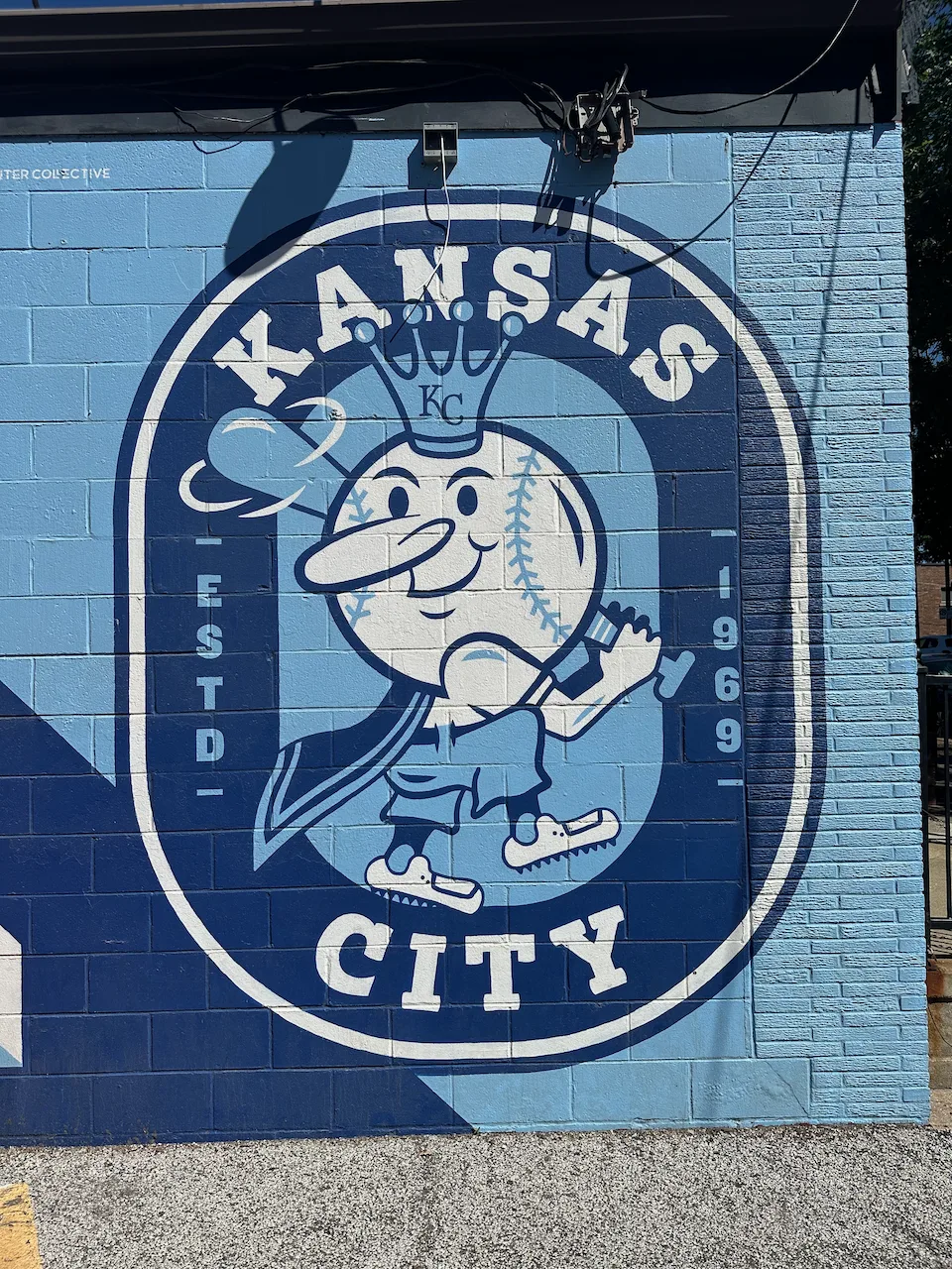 A cartoon baseball character with a crown is painted on a blue brick wall, featuring the text KANSAS CITY and ESTD 1969.