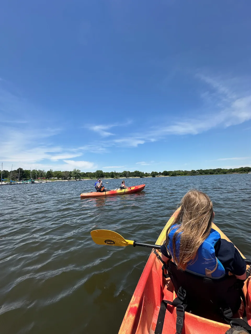 Two people in a red kayak paddle on a calm lake under a clear blue sky, with another kayak visible in the background.