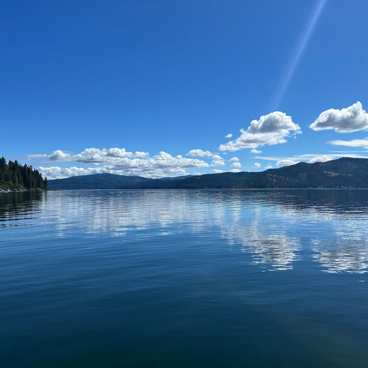 A calm lake reflects the clouds and sky, surrounded by forested hills under a bright blue sky.