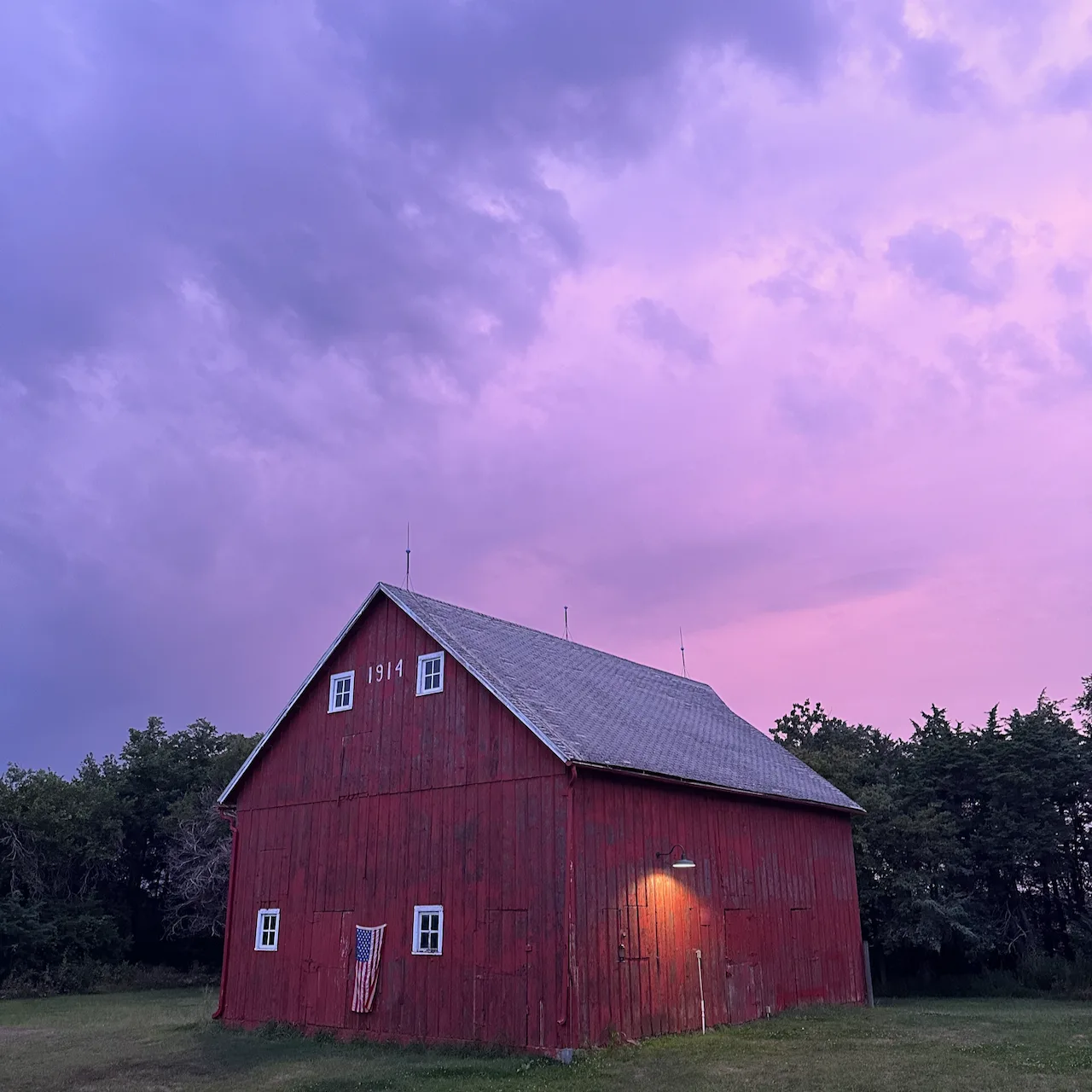 A red barn with the year 1914 painted on it stands under a dramatic evening sky with a purple hue.