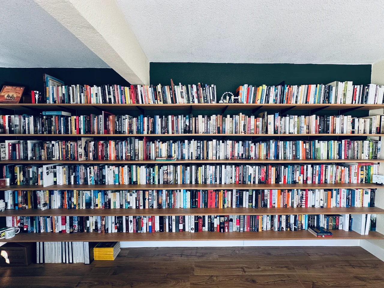 A large bookshelf filled with a diverse collection of books lines the wall of a room with wooden flooring.