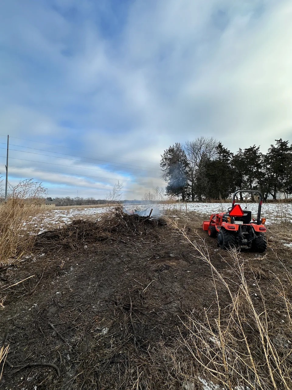 A red tractor sits beside a partially burned pile of debris in a grassy, winter landscape under a cloudy sky.