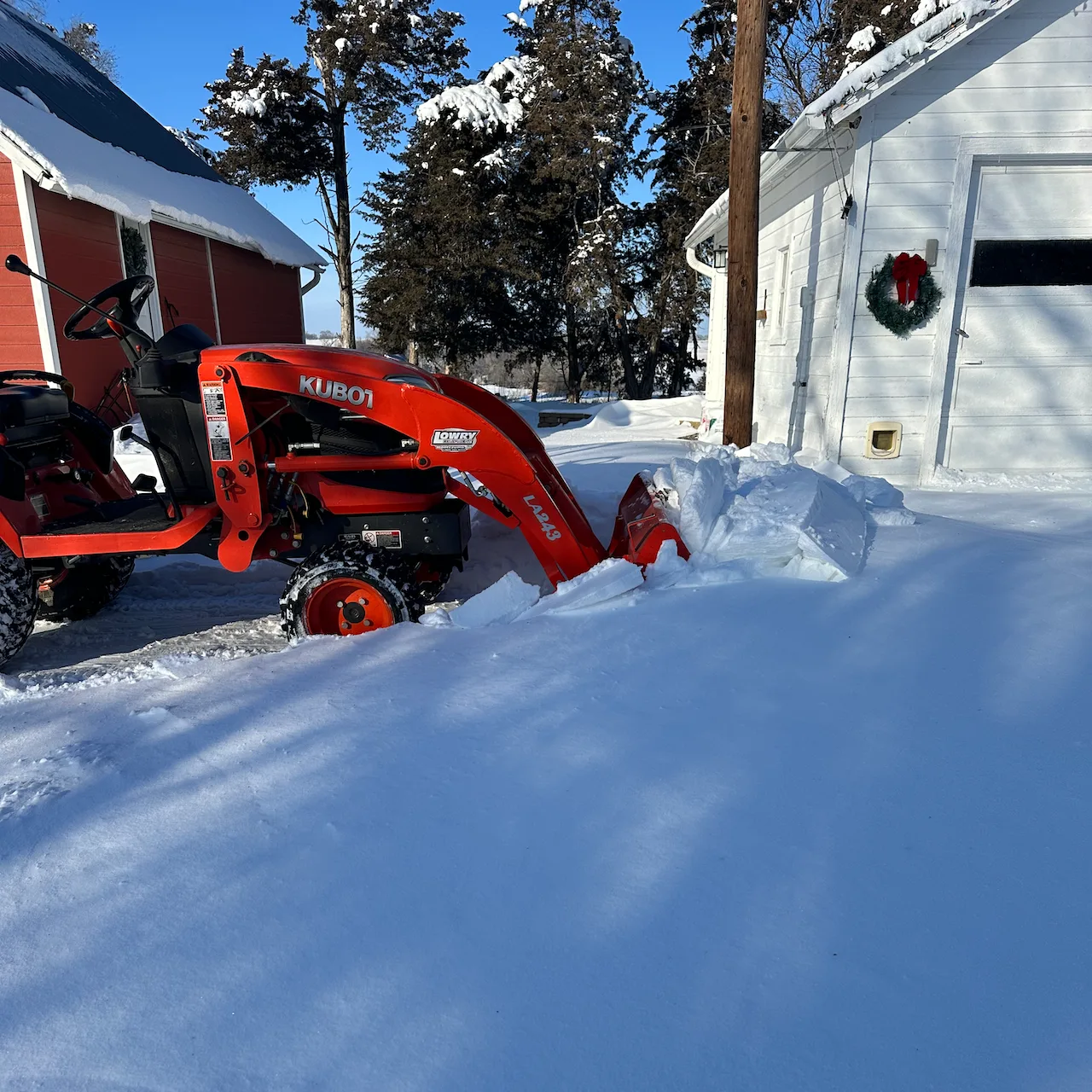 A snowplow attached to a small vehicle is clearing snow near a building decorated with a wreath.