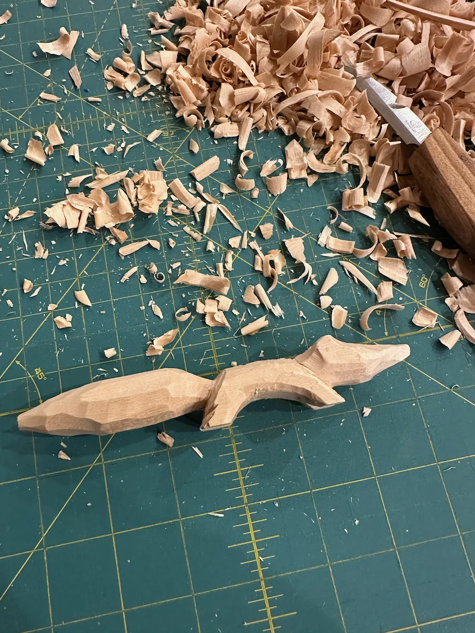 A partially carved wooden object is surrounded by wood shavings on a cutting mat, with a carving tool nearby.