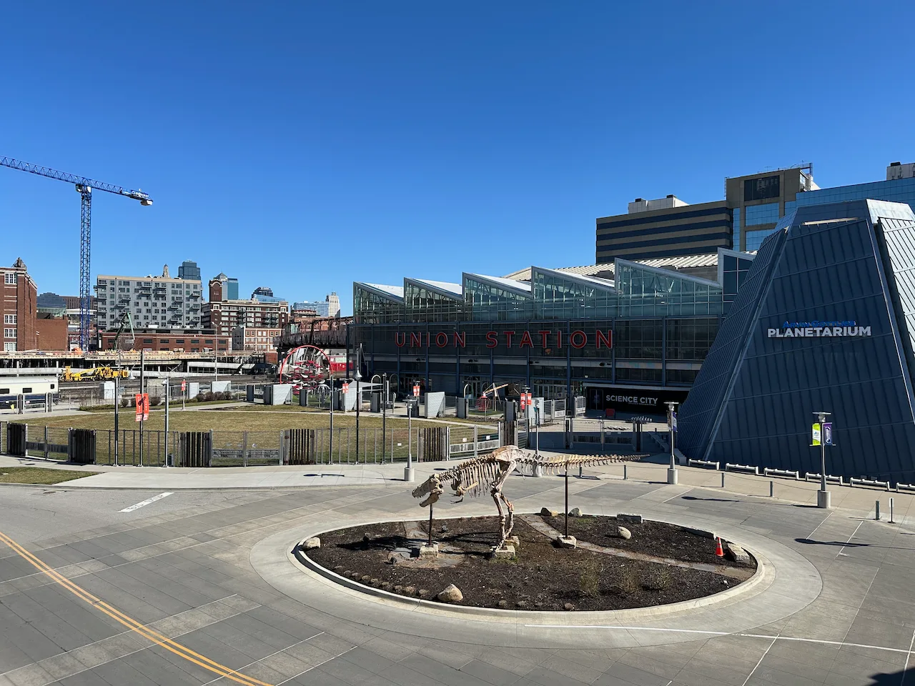 A cityscape features a dinosaur skeleton display, a modern building labeled PLANETARIUM, and a train station marked UNION STATION, with a construction crane in the background under a clear blue sky.