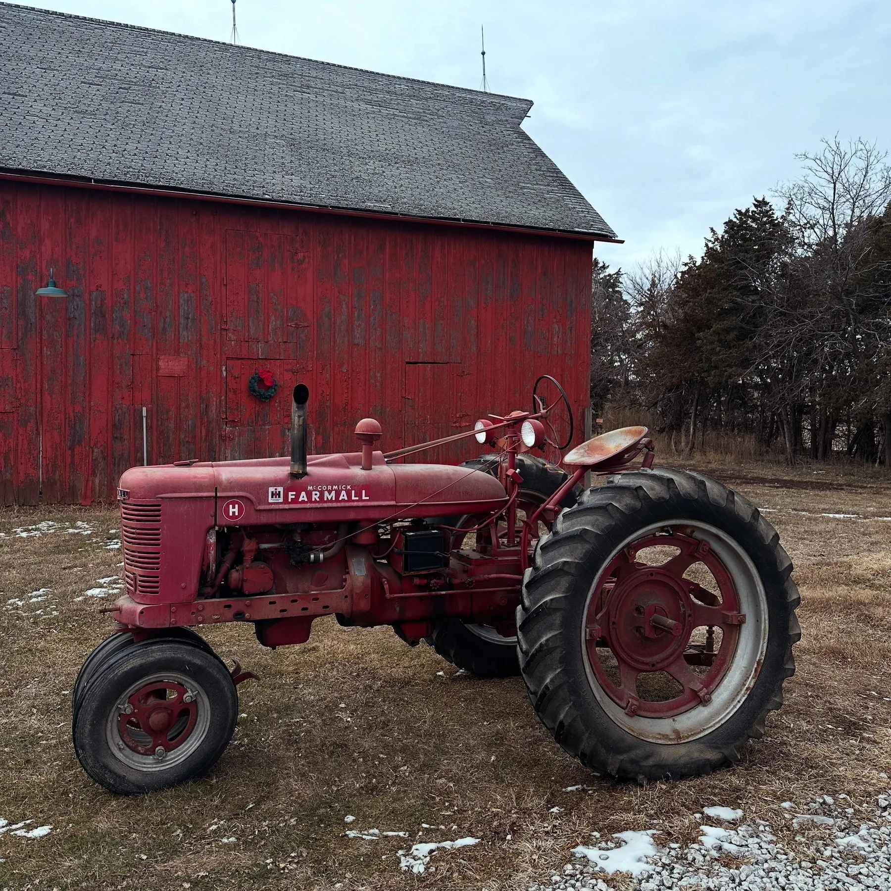 A red Farmall tractor is parked in front of an old, weathered red barn surrounded by bare trees and patches of snow on the ground.