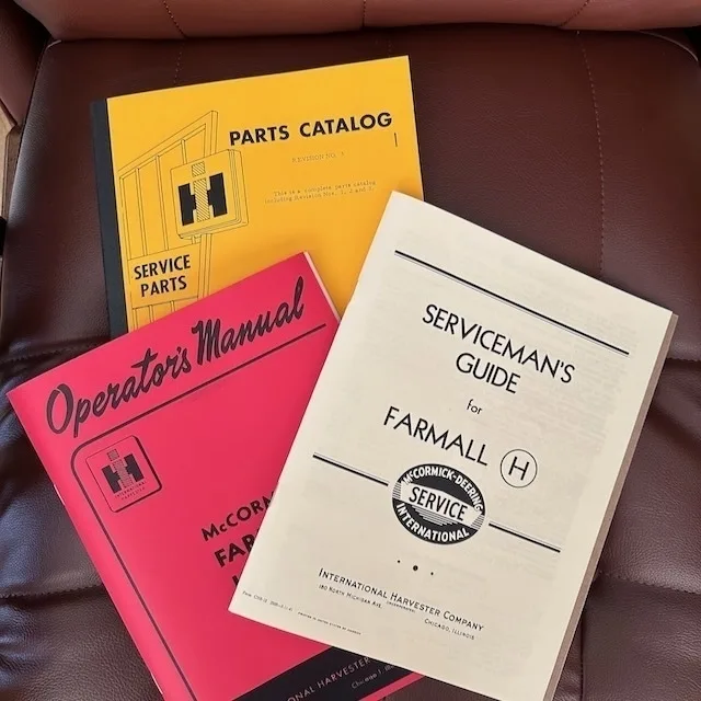 Three vintage manuals for Farmall tractors, including a parts catalog, operator's manual, and serviceman's guide, are displayed on a leather chair.
