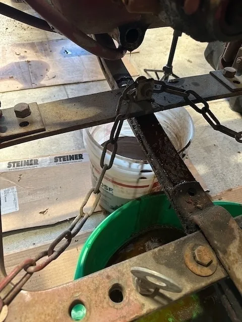 A chain is attached to a rusty metal frame above a green container, with cardboard and a white bucket in the background.