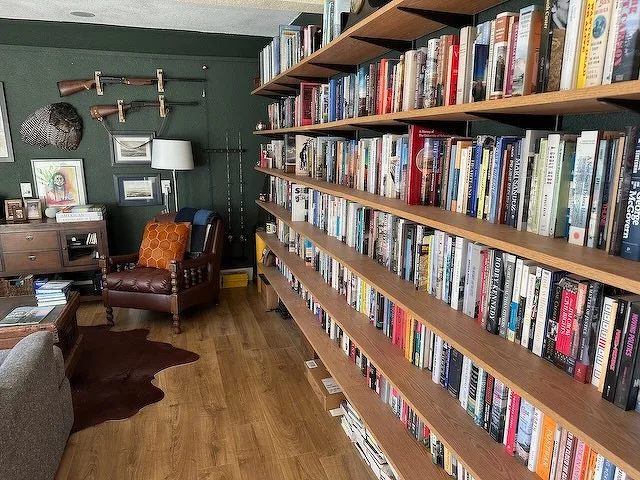 A cozy room features a large wall of bookshelves filled with books, a leather armchair with a pillow, and wall-mounted decorative items like rifles and framed art.