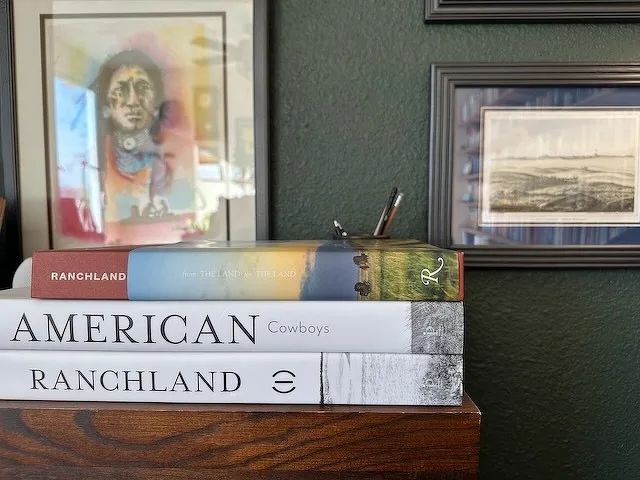 A stack of books with titles related to American landscapes and cowboy culture is positioned on top of a wooden surface, accompanied by framed art and photographs in the background.