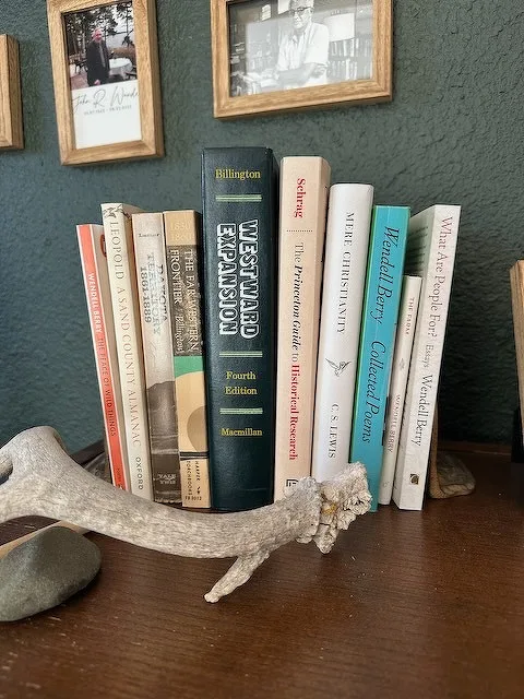 A bookshelf displays a collection of books alongside an antler and a small stone, with framed photos on the wall behind.