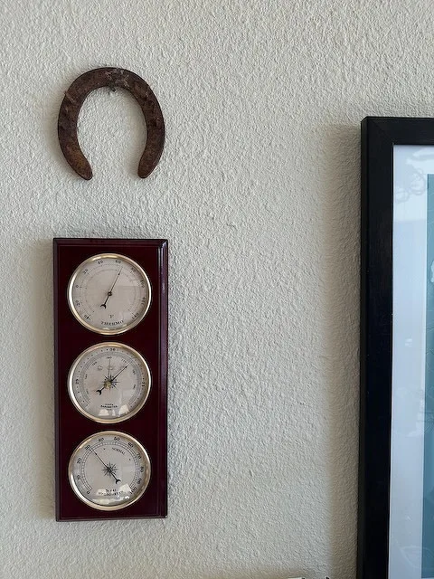 A wooden plaque with three round meteorological instruments is mounted on a wall beneath a hanging horseshoe, next to a framed picture.